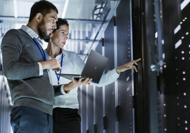Man holding a laptop and woman in a dark server room pointing at a server machine